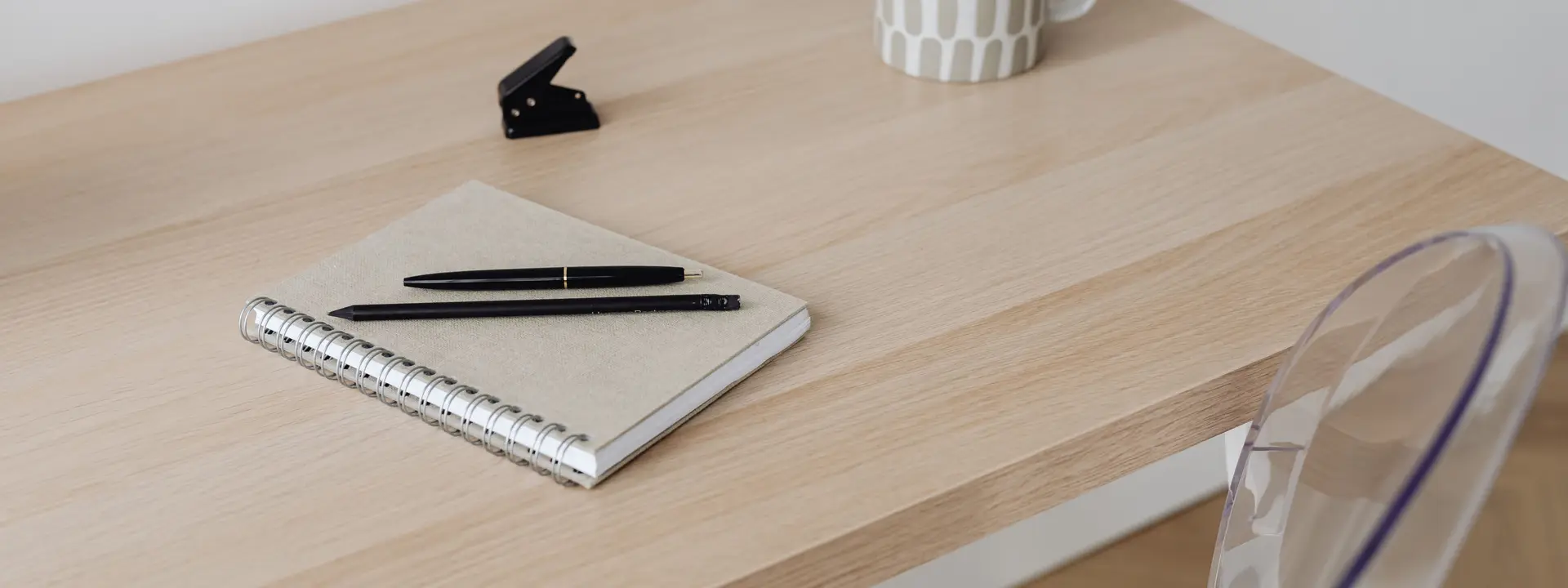 A notebook, pens and a mug on a light wooden desk